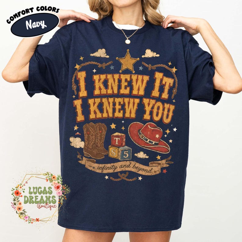 May include: A navy blue t-shirt featuring a graphic print with the text I KNEW IT I KNEW YOU and infinity and beyond. The design includes a gold star, a pair of brown cowboy boots, a red cowboy hat, and two alphabet blocks labelled T and S 5.