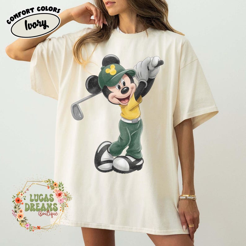 May include: Ivory-colored t-shirt featuring a cartoon Mickey Mouse playing golf. Mickey is wearing a green hat and pants, a yellow shirt, and white gloves. The shirt has the words "COMFORT COLORS" and "Ivory" printed on it. The bottom of the shirt has the words "LUCAS DREAMS BOUTIQUE".