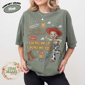 May include: A moss green t-shirt featuring a graphic of a cowgirl character, a red hat, brown cowboy boots, and alphabet blocks. The text reads: You've got a friend in me, infinity and beyond, and I knew it I knew you. A comfort colors label is visible.