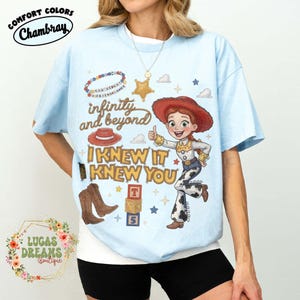 May include: A light blue t-shirt featuring a graphic print of a cowgirl character, a red cowboy hat, brown boots, alphabet blocks, and a beaded necklace. The text reads "infinity and beyond" and "I KNEW IT KNEW YOU" in stylized yellow and brown lettering.
