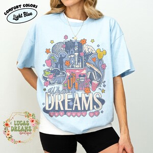 K&ouml;nnte beinhalten: Hellblaues T-Shirt mit einem farbenfrohen Grafikdesign mit einem Schloss, einer Einschienenbahn und dem Text "What Dreams Are Made Of". Das Shirt hat auch den Text "Comfort Colors" und "Light Blue" in einem Kreis.