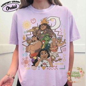 May include: A light purple t-shirt featuring a graphic print of characters from the movie Moana, including Moana, Maui, Pua, Heihei, and Gramma Tala. The design includes a large white fishhook, various colorful tribal patterns, and symbols.