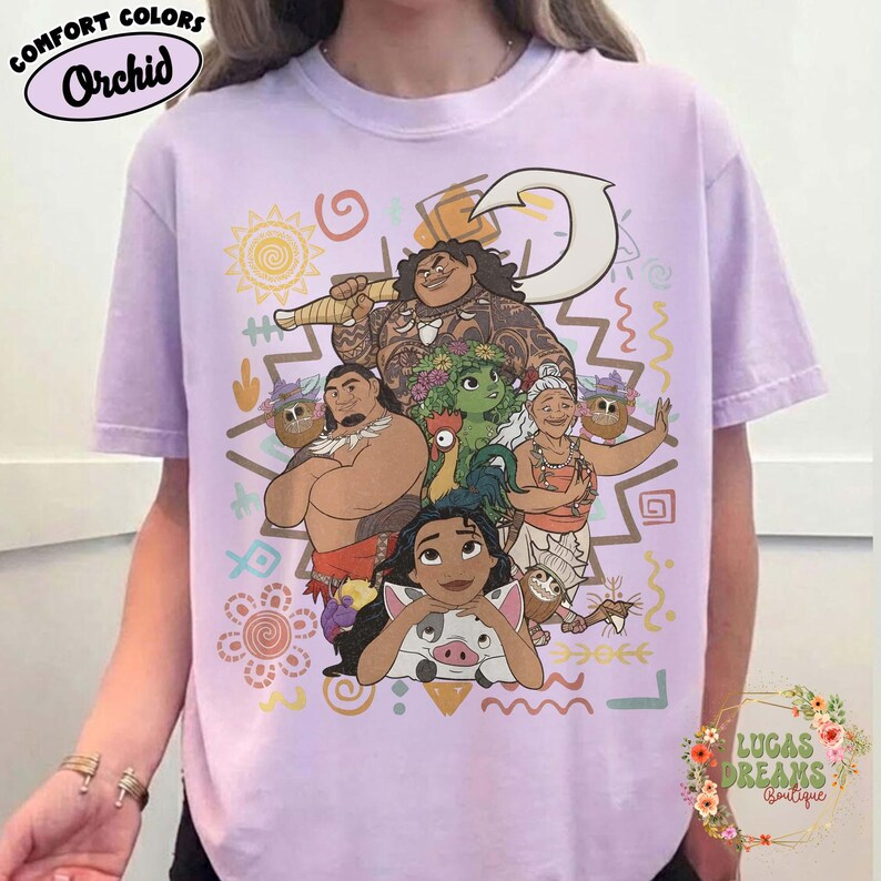 May include: A light purple t-shirt featuring a graphic print of characters from the movie Moana, including Moana, Maui, Pua, Heihei, and Gramma Tala. The design includes a large white fishhook, various colorful tribal patterns, and symbols.