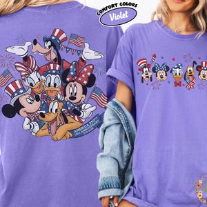 以下が含まれることがあります： 紫色のComfort Colors Tシャツ。ミッキー、ミニー、ドナルド、グーフィーなど、愛国的な衣装を着たディズニーのキャラクターが、アメリカの国旗と花火とともに描かれています。Tシャツの背面には、グラフィックの大きなバージョンがあります。「America 250th USA Anniversary 1776-2026」と書かれています。