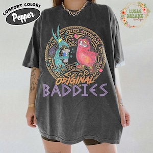 Puede incluir: Camiseta gris oscuro con un diseño circular que presenta personajes de dibujos animados en turquesa y rosa, con el texto "ORIGINAL BADDIES" en morado. La camiseta tiene un aspecto vintage y desgastado.