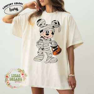 May include: Ivory-colored t-shirt featuring a Halloween-themed graphic of Mickey Mouse dressed as a mummy, holding a jack-o'-lantern. The shirt has the words "Comfort Colors" and "Ivory" printed on it. The shirt is from Lucas Dreams Boutique.