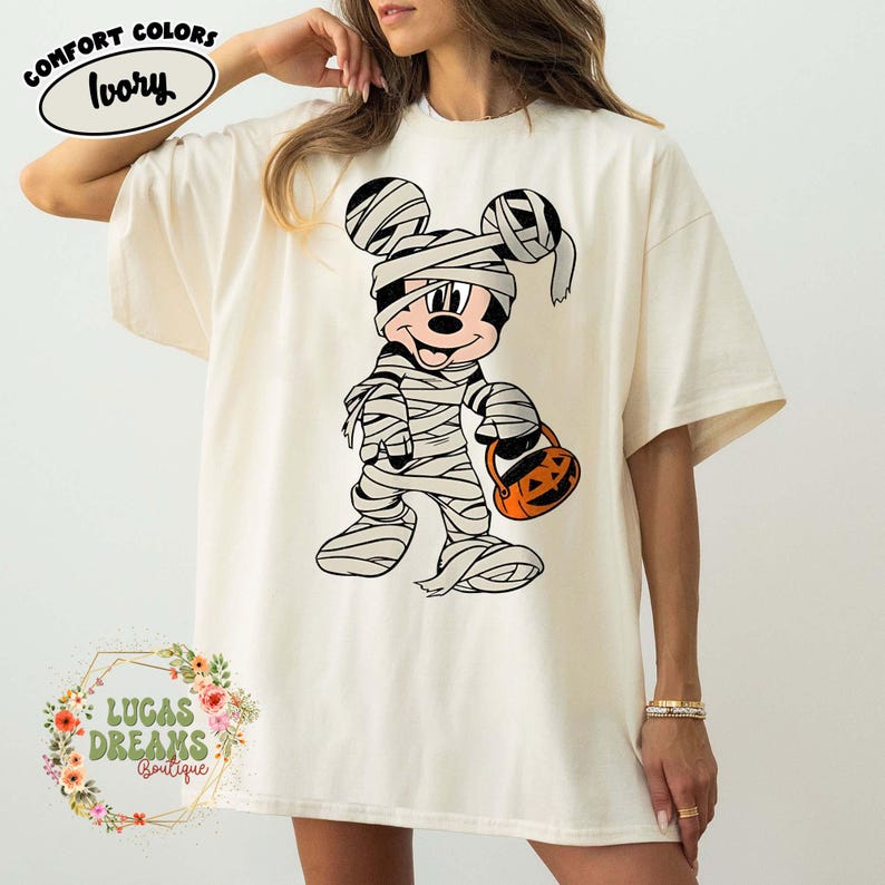 May include: Ivory-colored t-shirt featuring a Halloween-themed graphic of Mickey Mouse dressed as a mummy, holding a jack-o'-lantern. The shirt has the words "Comfort Colors" and "Ivory" printed on it. The shirt is from Lucas Dreams Boutique.