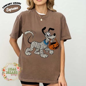 May include: Brown t-shirt with a cartoon dog wrapped in bandages, holding an orange jack-o'-lantern bucket. The shirt has the words "Comfort Colors Espresso" in a circular design at the top. The bottom left corner has the text "Lucas Dreams Boutique".