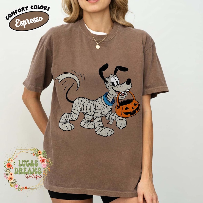 May include: Brown t-shirt with a cartoon dog wrapped in bandages, holding an orange jack-o'-lantern bucket. The shirt has the words "Comfort Colors Espresso" in a circular design at the top. The bottom left corner has the text "Lucas Dreams Boutique".