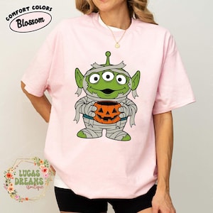May include: Pink t-shirt with a cartoon alien mummy holding a jack-o'-lantern. The alien is green with three eyes and wrapped in bandages. The shirt has the text "COMFORT COLORS Blossom" at the top.