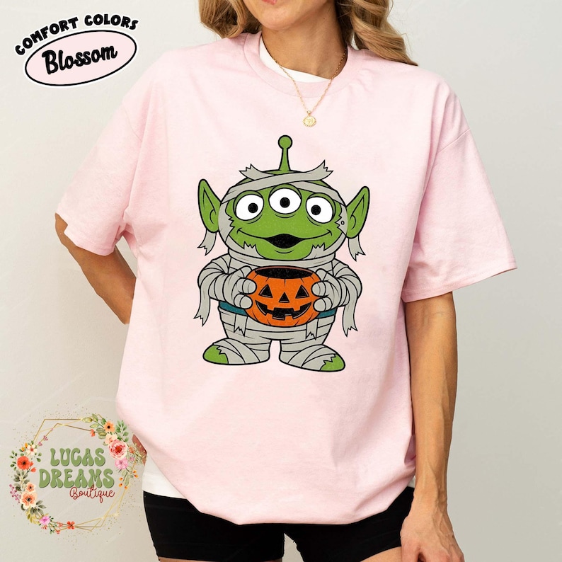 May include: Pink t-shirt with a cartoon alien mummy holding a jack-o'-lantern. The alien is green with three eyes and wrapped in bandages. The shirt has the text "COMFORT COLORS Blossom" at the top.