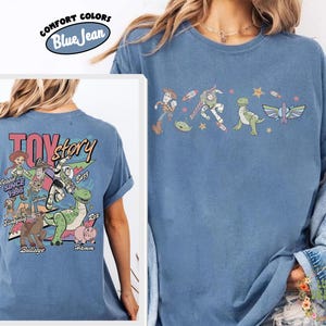 May include: Blue jean Comfort Colors t-shirt featuring Toy Story characters. The front displays a line of characters including Woody and Buzz Lightyear. The back of the shirt has the Toy Story logo and other characters like Jessie and Rex.