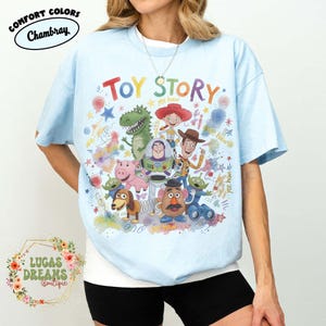 Pu&ograve; includere: T-shirt azzurra con la scritta "TOY STORY" a colori e personaggi dei cartoni animati. La maglietta ha le parole "Comfort Colors Chambray" stampate in alto a sinistra e "LUCAS DREAMS Boutique" in basso a sinistra.