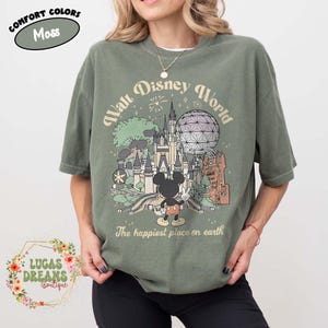 May include: Moss green t-shirt featuring a Walt Disney World graphic with Mickey Mouse. The design includes a castle, Spaceship Earth, and the text "The happiest place on earth". The shirt has short sleeves and a relaxed fit.