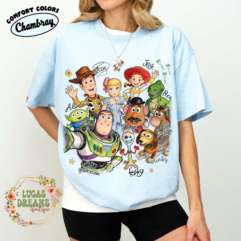 Op de afbeelding: Een lichtblauw t-shirt met een kleurrijke grafische print van Toy Story-personages, waaronder Woody, Buzz Lightyear, Bo Peep, Jessie, Rex, Mr. Potato Head, Slinky Dog, Forky en een Alien, met hun namen in een cursief handschrift.