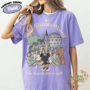 May include: Violet Comfort Colors t-shirt featuring a Disney World graphic. The design includes a castle, Spaceship Earth, and Mickey Mouse. Text reads "Disney World" and "The happiest place on earth."