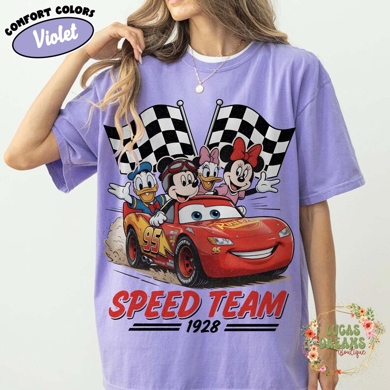 Op de afbeelding: Violet Comfort Colors T-shirt met een afbeelding van Disney-personages Mickey Mouse, Donald Duck, Daisy Duck en Minnie Mouse in een rode raceauto. De woorden "SPEED TEAM 1928" staan eronder gedrukt, met twee geblokte vlaggen op de achtergrond.