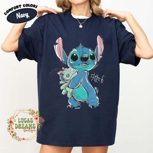 May include: A navy blue short-sleeve t-shirt featuring a graphic illustration of the character Stitch holding a small doll. The design includes the text Stitch in a stylized font. A circular label in the corner reads Comfort Colors Navy.
