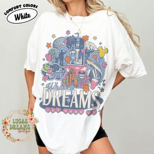 K&ouml;nnte beinhalten: Wei&szlig;es T-Shirt mit einem farbenfrohen Grafikdesign mit einem Schloss, einer Silhouette einer Person und dem Text "What Dreams Are Made Of". Das Shirt hat auch den Text "Comfort Colors White" und "Lucas Dreams Boutique."