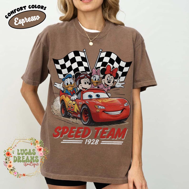 Op de afbeelding: Bruin t-shirt met een afbeelding van Disney-personages in een rode raceauto, geblokte vlaggen en de woorden "Speed Team 1928". Het shirt heeft ook de woorden "Comfort Colors Espresso" in een ovaal.