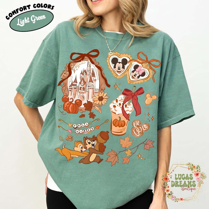 May include: Light green t-shirt with a fall-themed graphic. The design includes a castle, pumpkins, Mickey and Minnie Mouse, a pumpkin spice latte, Chip and Dale, and the words "Fall Vibes."