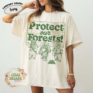 Puede incluir: Camiseta color marfil con un gráfico verde que dice "Protect our Forests!" y presenta tres Ewoks. El logo de Star Wars está en la parte inferior. La camiseta es de Lucas Dreams Boutique.