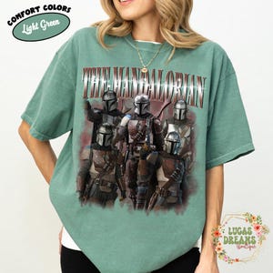 Puede incluir: Una camiseta de manga corta de color verde claro con un estampado gr&aacute;fico de cinco personajes mandalorianos con armadura plateada. El texto THE MANDALORIAN est&aacute; impreso en una fuente met&aacute;lica llamativa sobre los personajes. Se ve una etiqueta.