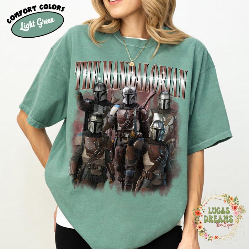 Puede incluir: Una camiseta de manga corta de color verde claro con un estampado gr&aacute;fico de cinco personajes mandalorianos con armadura plateada. El texto THE MANDALORIAN est&aacute; impreso en una fuente met&aacute;lica llamativa sobre los personajes. Se ve una etiqueta.