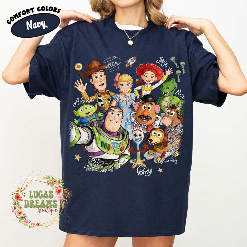 Op de afbeelding: Een marineblauw t-shirt met korte mouwen en een kleurrijke grafische print van diverse animatiefiguren, waaronder Woody, Buzz Lightyear, Jessie, Bo Peep, Rex, Mr. Potato Head, Slinky Dog, Bullseye en Forky, met hun namen in cursief schrift.