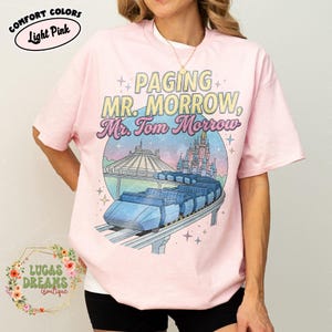 May include: Pale pink t-shirt with a graphic design. The design includes the text "PAGING MR. MORROW, Mr. Tom Morrow", a futuristic monorail, and a castle. The shirt also has the words "COMFORT COLORS" and "Light Pink".