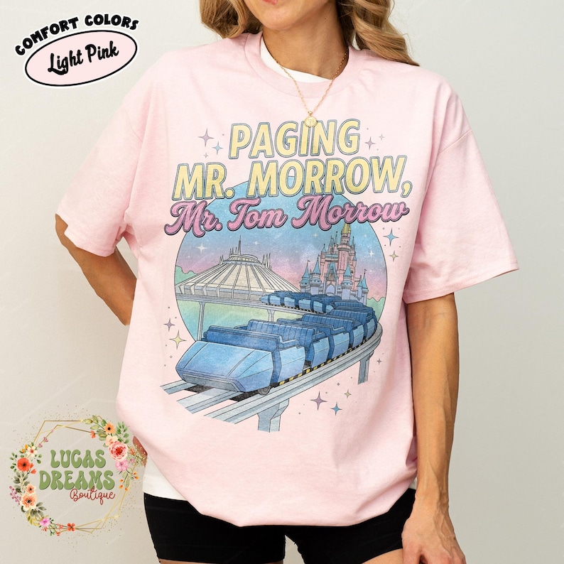 May include: Pale pink t-shirt with a graphic design. The design includes the text "PAGING MR. MORROW, Mr. Tom Morrow", a futuristic monorail, and a castle. The shirt also has the words "COMFORT COLORS" and "Light Pink".