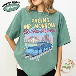 May include: Light green t-shirt featuring the text "PAGING MR. MORROW, Mr. Tom Morrow" and a graphic of a futuristic train, Space Mountain, and a castle. The shirt has the words "COMFORT COLORS" and "Light Green" on it.
