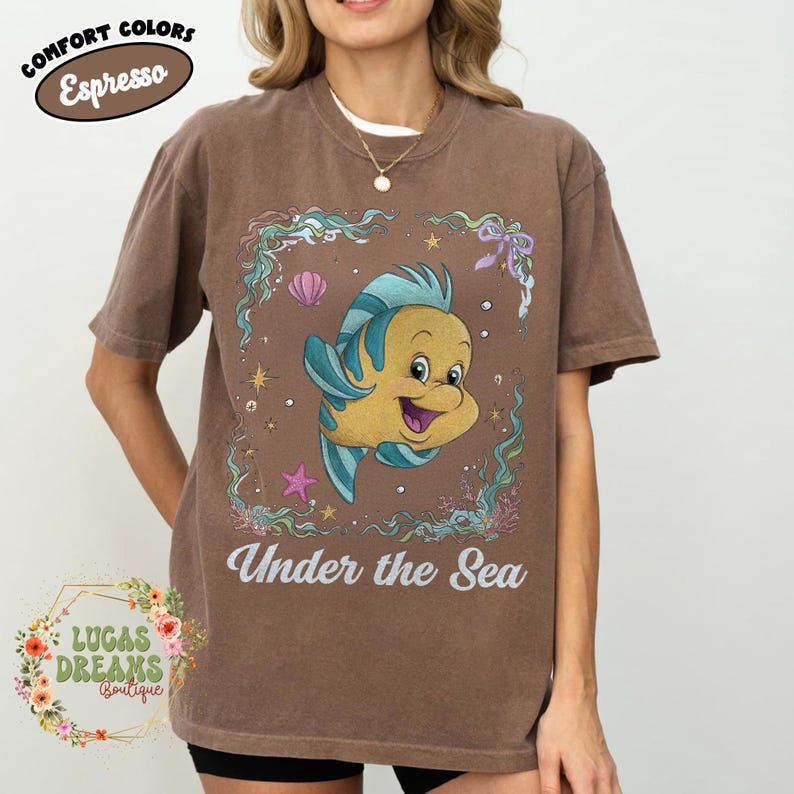 May include: Espresso-colored t-shirt featuring a cartoon fish character with blue and yellow accents, surrounded by aquatic elements. The phrase "Under the Sea" is printed below the graphic. The shirt also has the text "Comfort Colors" and "Espresso".