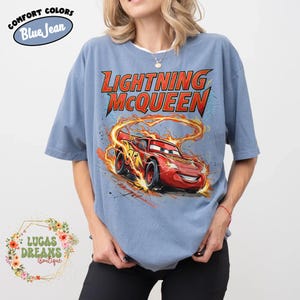 May include: Blue short-sleeve t-shirt featuring a graphic of Lightning McQueen from the movie Cars. The design includes the text "LIGHTNING McQUEEN" in red and yellow, with the character surrounded by flames. The shirt has the text "COMFORT COLORS Blue Jean" on the top left.