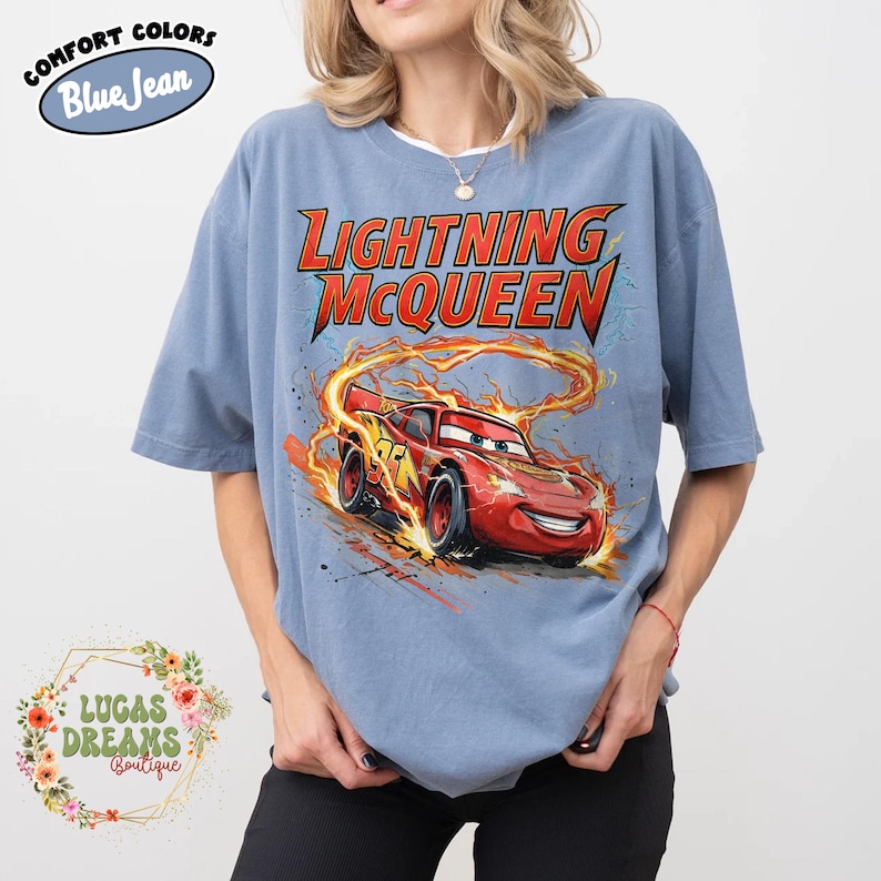 May include: Blue short-sleeve t-shirt featuring a graphic of Lightning McQueen from the movie Cars. The design includes the text "LIGHTNING McQUEEN" in red and yellow, with the character surrounded by flames. The shirt has the text "COMFORT COLORS Blue Jean" on the top left.