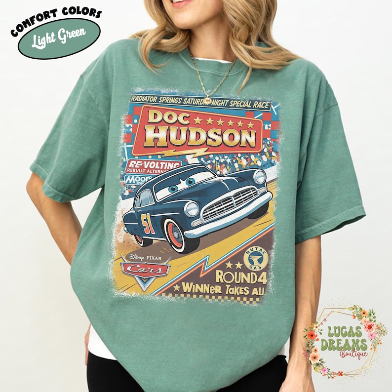 May include: Light green t-shirt featuring a vintage-style graphic of Doc Hudson from the Disney Pixar Cars movie. The design includes the text "Doc Hudson," "Round 4," and "Winner Takes All." The shirt is labeled "Comfort Colors."