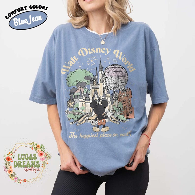 May include: Blue short-sleeved t-shirt with a graphic of Walt Disney World, including Mickey Mouse, a castle, and the text "The happiest place on earth." The shirt also has the text "Comfort Colors" and "Blue Jean."