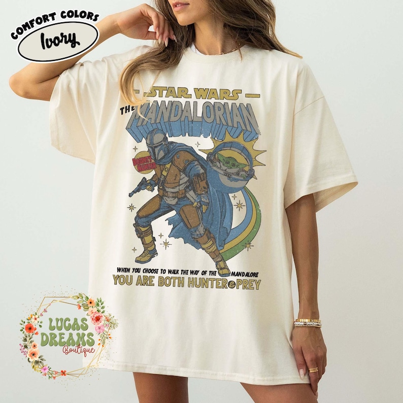 Puede incluir: Una camiseta color marfil con un estampado gr&aacute;fico de The Mandalorian y Grogu. El texto dice STAR WARS THE MANDALORIAN, WHEN YOU CHOOSE TO WALK THE WAY OF THE MANDALORE YOU ARE BOTH HUNTER & PREY. El dise&ntilde;o incluye una capa azul y estrellas.