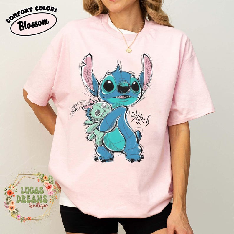 May include: A light pink t-shirt featuring a graphic illustration of the blue character Stitch holding a small doll. The design includes the text Stitch in a stylized font. The shirt has short sleeves and a crew neckline, with the Comfort Colors Blossom label.