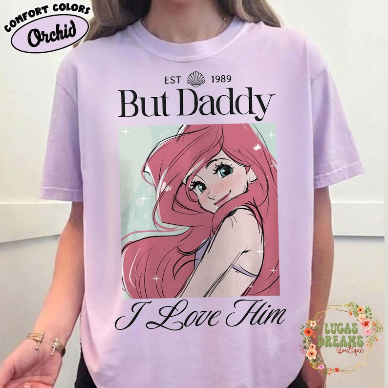 May include: Orchid-colored t-shirt with the text "But Daddy I Love Him" and an illustration of a cartoon character with pink hair. The shirt also has the text "EST 1989" above the illustration.