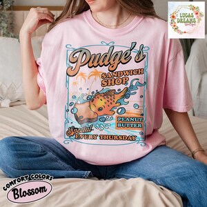 Pu&ograve; includere: T-shirt rosa chiaro con grafica "Pudge's Sandwich Shop". Il design presenta un pesce dei cartoni animati, palme e le parole "Peanut Butter Special Every Thursday". La maglietta &egrave; Comfort Colors Blossom.