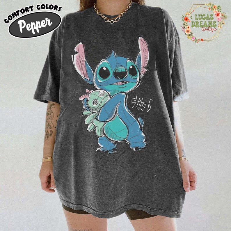 May include: A charcoal gray short-sleeve t-shirt featuring a graphic of the blue character Stitch holding a small doll. The design includes the text Stitch in a stylized script. The shirt has a vintage-washed appearance and a crew neckline.
