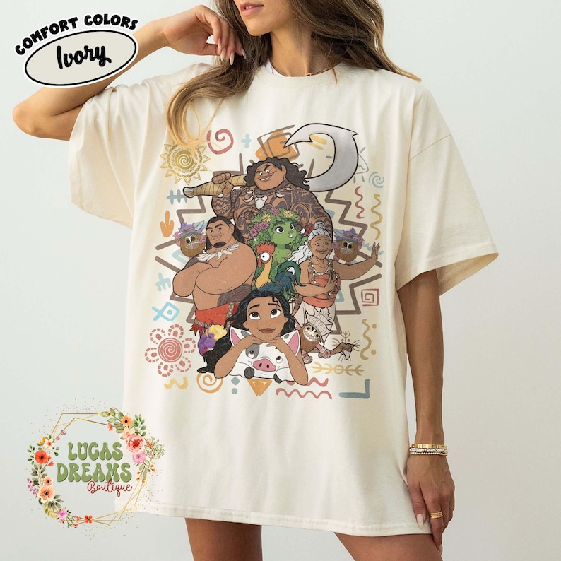 May include: An ivory short-sleeve t-shirt featuring a graphic print of characters from the movie Moana, including Moana, Maui, Pua, Heihei, and Gramma Tala. The design includes tribal-inspired patterns, a hook, and various colorful symbols.