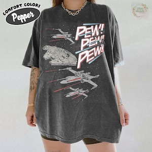 May include: Dark grey t-shirt featuring a space battle design with the text "PEW! PEW! PEW!". The graphic includes spaceships and a starfield. The shirt has a vintage, worn look and the words "Comfort Colors" and "Pepper" are printed on the top left.