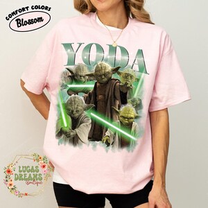Puede incluir: Una camiseta de manga corta de color rosa claro con un estampado gr&aacute;fico del personaje Yoda en varias poses con sables de luz verdes. La palabra YODA est&aacute; impresa en letras grandes sobre el personaje. Incluye una etiqueta Comfort Colors Blossom.