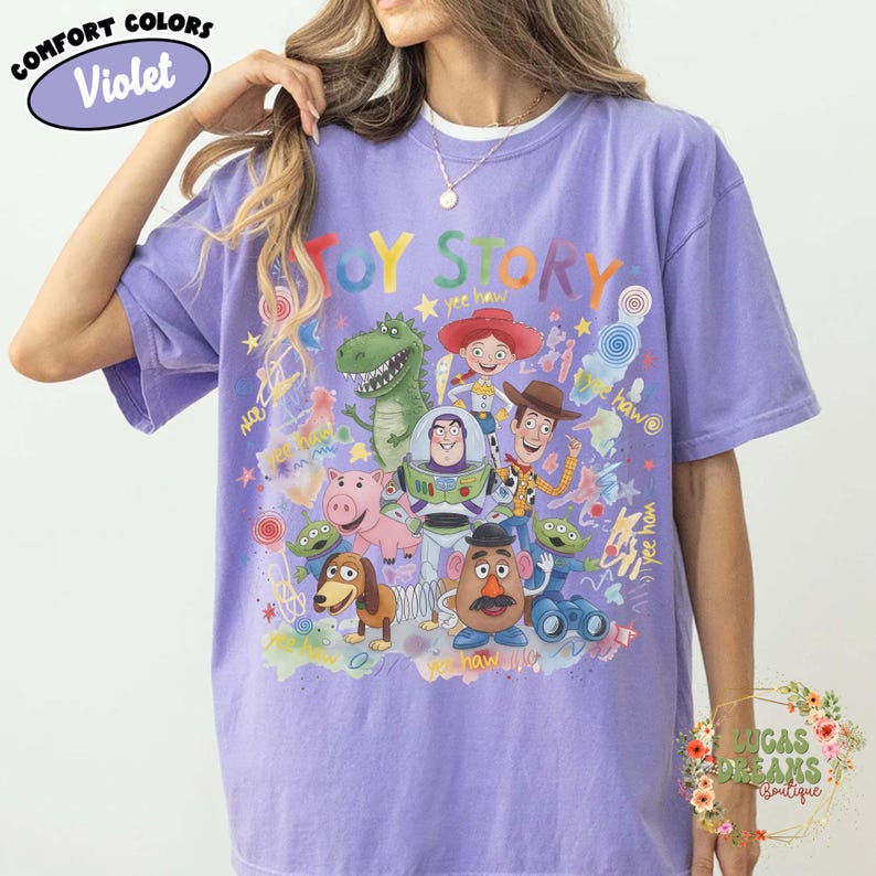 Pu&ograve; includere: T-shirt Comfort Colors viola con una grafica colorata dei personaggi di Toy Story. Il design include Woody, Buzz Lightyear e altri personaggi del film d'animazione. La maglietta presenta la scritta "Toy Story" in un font arcobaleno.