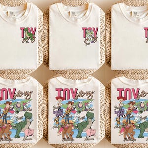 Peut inclure: T-shirts blanc cass&eacute; avec le texte "Toy Story" en rose et noir. Certains t-shirts ont un petit dessin de personnage, tandis que d'autres ont un dessin plus grand avec plusieurs personnages. Les t-shirts sont pr&eacute;sent&eacute;s sur une surface tiss&eacute;e.