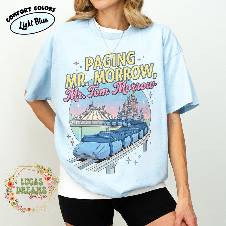 May include: Pale blue t-shirt featuring the text "PAGING MR. MORROW, Mr. Tom Morrow" and a graphic of a monorail and a castle. The shirt also has the words "COMFORT COLORS" and "Light Blue" on it.