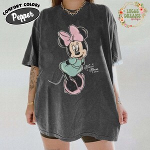 May include: A charcoal grey short-sleeve t-shirt featuring a graphic of Minnie Mouse wearing a pink bow and a teal dress. The text "Minnie Mouse xoxo" is printed in white script next to the character. The shirt has a vintage washed appearance.