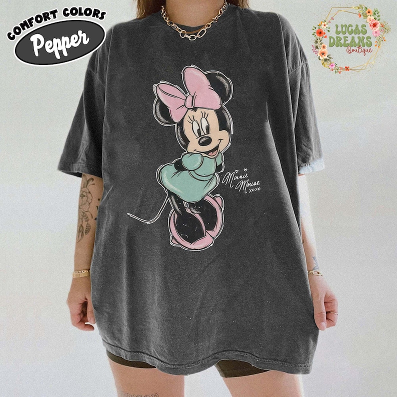 May include: A charcoal grey short-sleeve t-shirt featuring a graphic of Minnie Mouse wearing a pink bow and a teal dress. The text "Minnie Mouse xoxo" is printed in white script next to the character. The shirt has a vintage washed appearance.